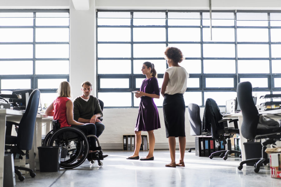 A photo of business team communicating at workplace. Female sitting on wheelchair in meeting. They are in brightly lit n creative office.